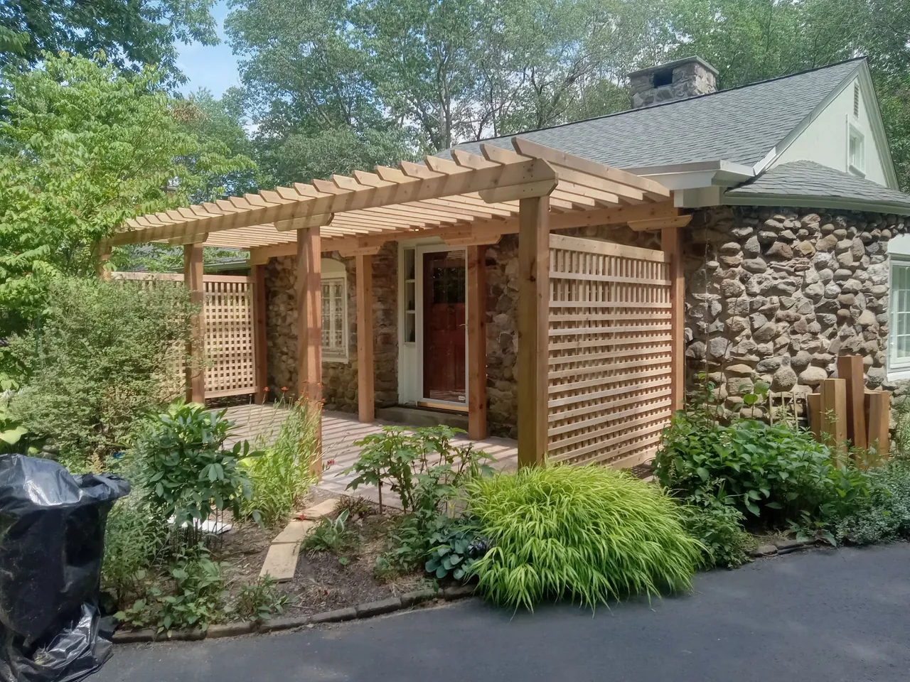 Wooden pergola over a stone house entrance with lattice and surrounding greenery